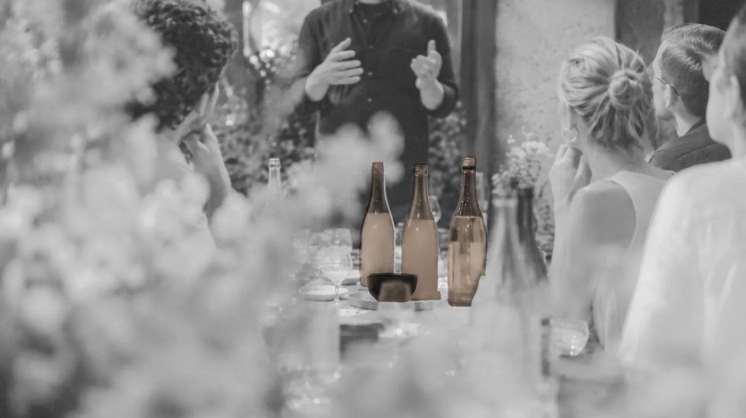 A black-and-white scene of a dinner table in Berlin — the founder gesturing at the head of the table, bottles and guests around it