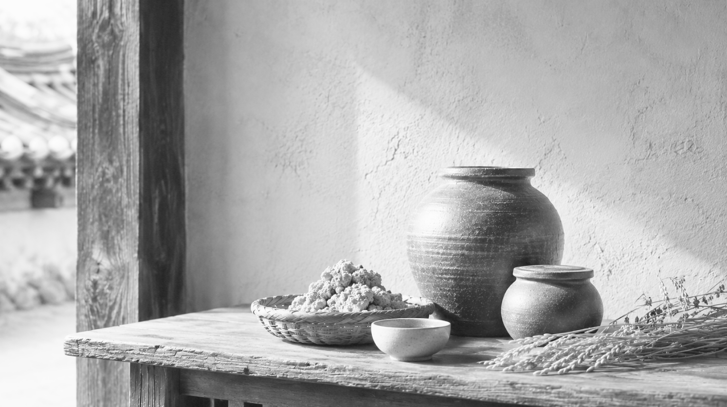 A black-and-white hanok scene — a suldok brewing jar, a nuruk basket, a cup and dried rice on a wooden table